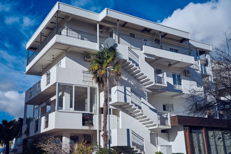 Immeuble résidentiel blanc à plusieurs étages avec balcons, escaliers extérieurs en métal, un palmier au premier plan, une antenne parabolique et des climatiseurs, sous un ciel bleu partiellement nuageux.