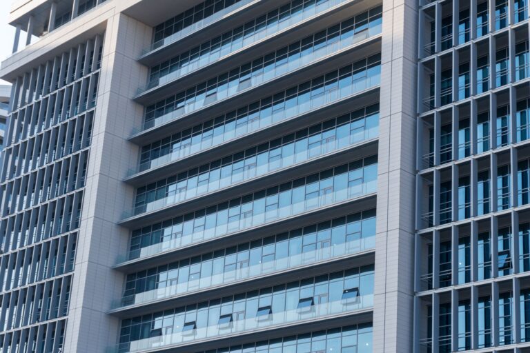 Façade d'un immeuble de bureaux moderne en verre et béton, avec des rangées de fenêtres et des balcons vitrés alternant avec des structures verticales en métal gris.