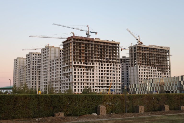Plusieurs immeubles résidentiels en béton en construction, à plusieurs étages, avec des grues à tour sous un ciel crépusculaire ; une haie verte et un bâtiment vitré à droite.