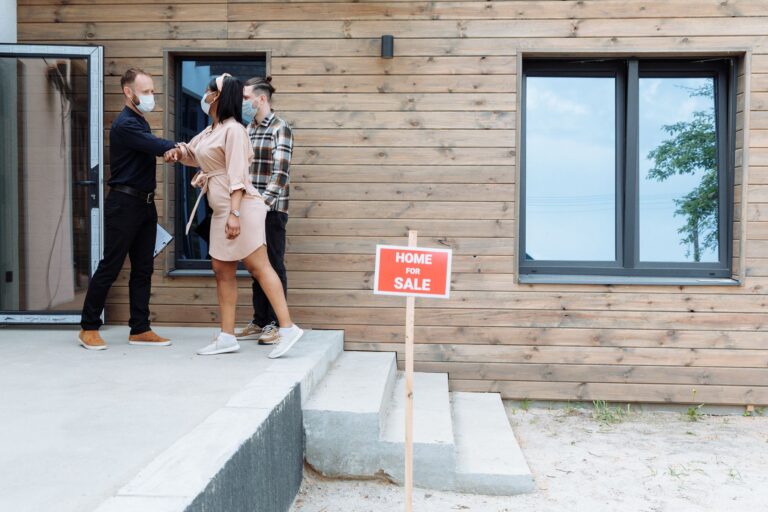 Un agent immobilier en chemise noire et masque accueille un couple, la femme en robe beige et l'homme en chemise à carreaux, devant une maison en bois avec un panneau rouge « HOME FOR SALE ».