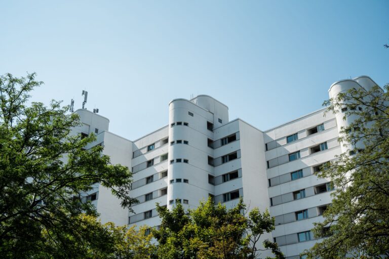 Vue en contre-plongée d'un immeuble résidentiel blanc aux balcons arrondis et fenêtres alignées, entouré d'arbres verts sous un ciel bleu dégagé, avec des antennes sur le toit.