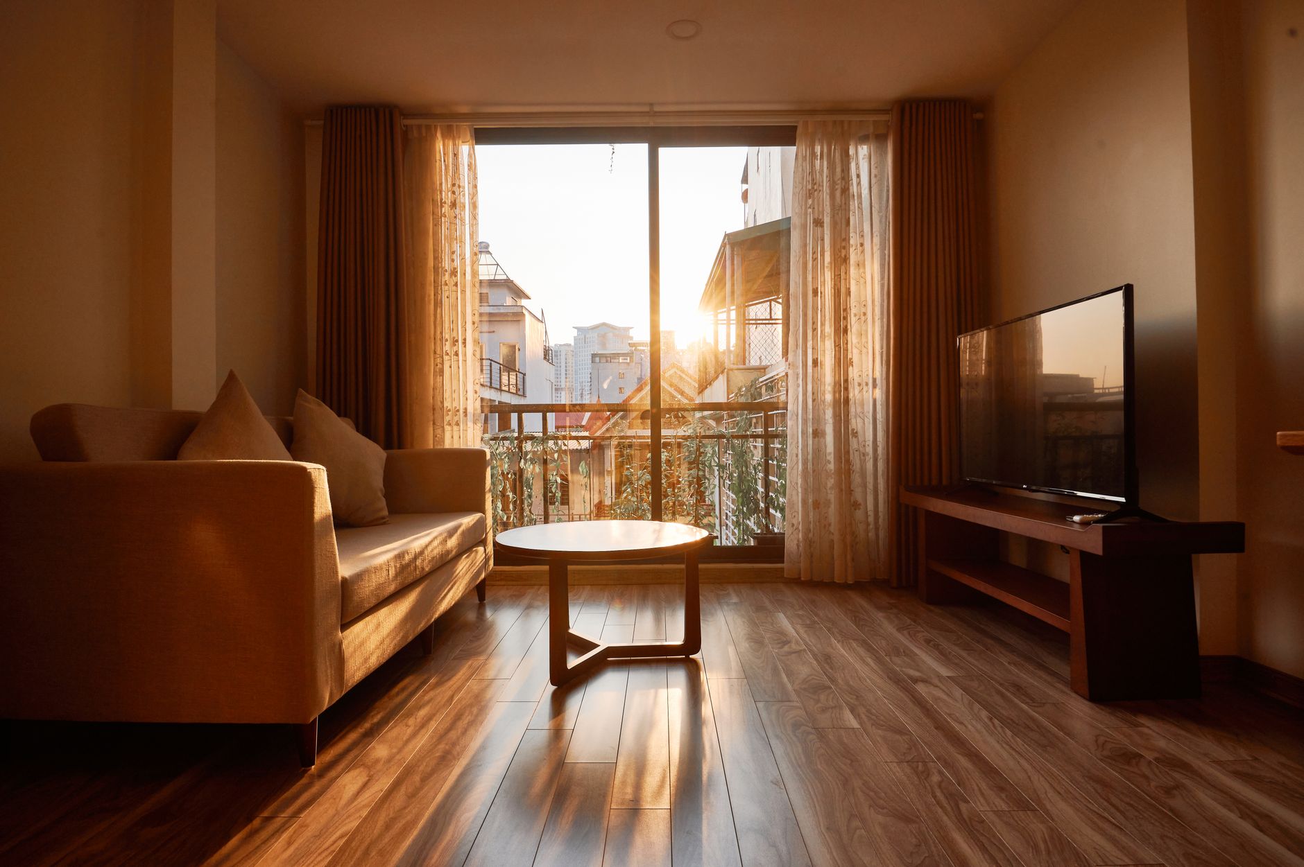 Salon meublé baigné de lumière dorée au coucher du soleil, avec un canapé beige et ses coussins, une table basse ronde en bois, un téléviseur sur un meuble TV en bois, et une baie vitrée aux rideaux donnant sur un balcon et des immeubles.