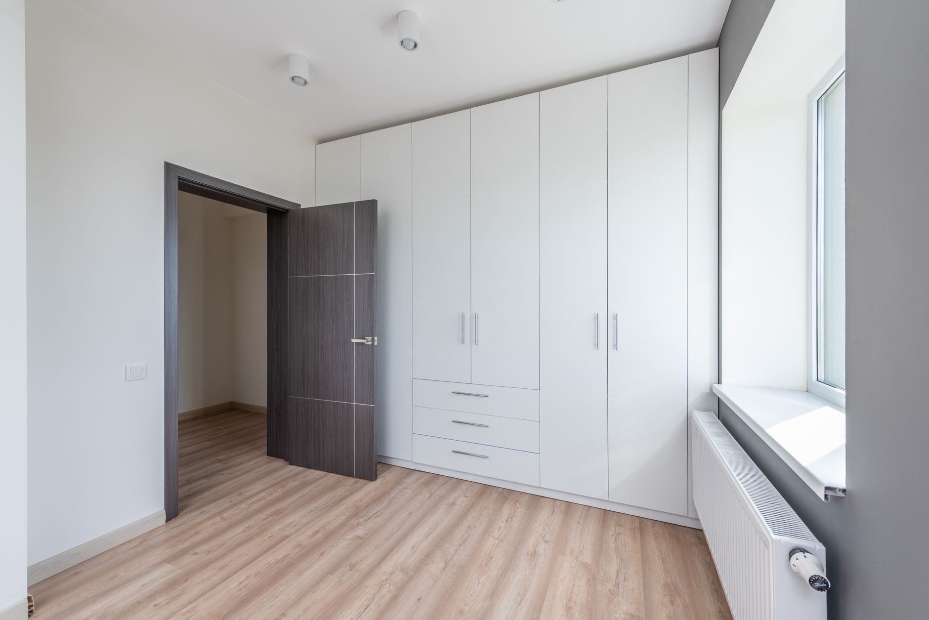 Pièce vide et lumineuse au parquet clair, avec une grande armoire blanche à portes et tiroirs, une porte en bois foncé ouverte, une fenêtre à rebord blanc et un radiateur.