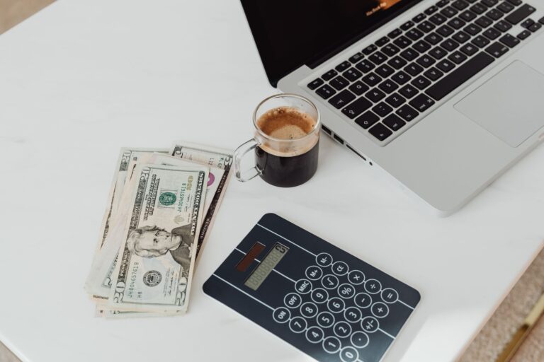 Table blanche avec ordinateur portable, calculatrice, tasse de café et pile de billets de dollars américains.