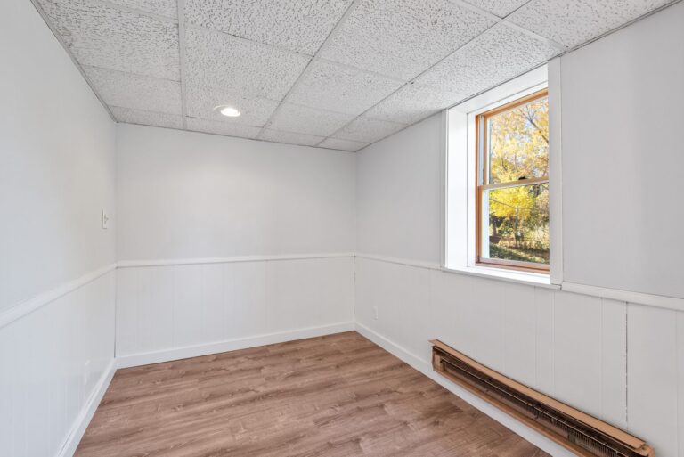 Pièce vide, murs blancs, plafond acoustique, parquet imitation bois, fenêtre sur paysage automnal.
