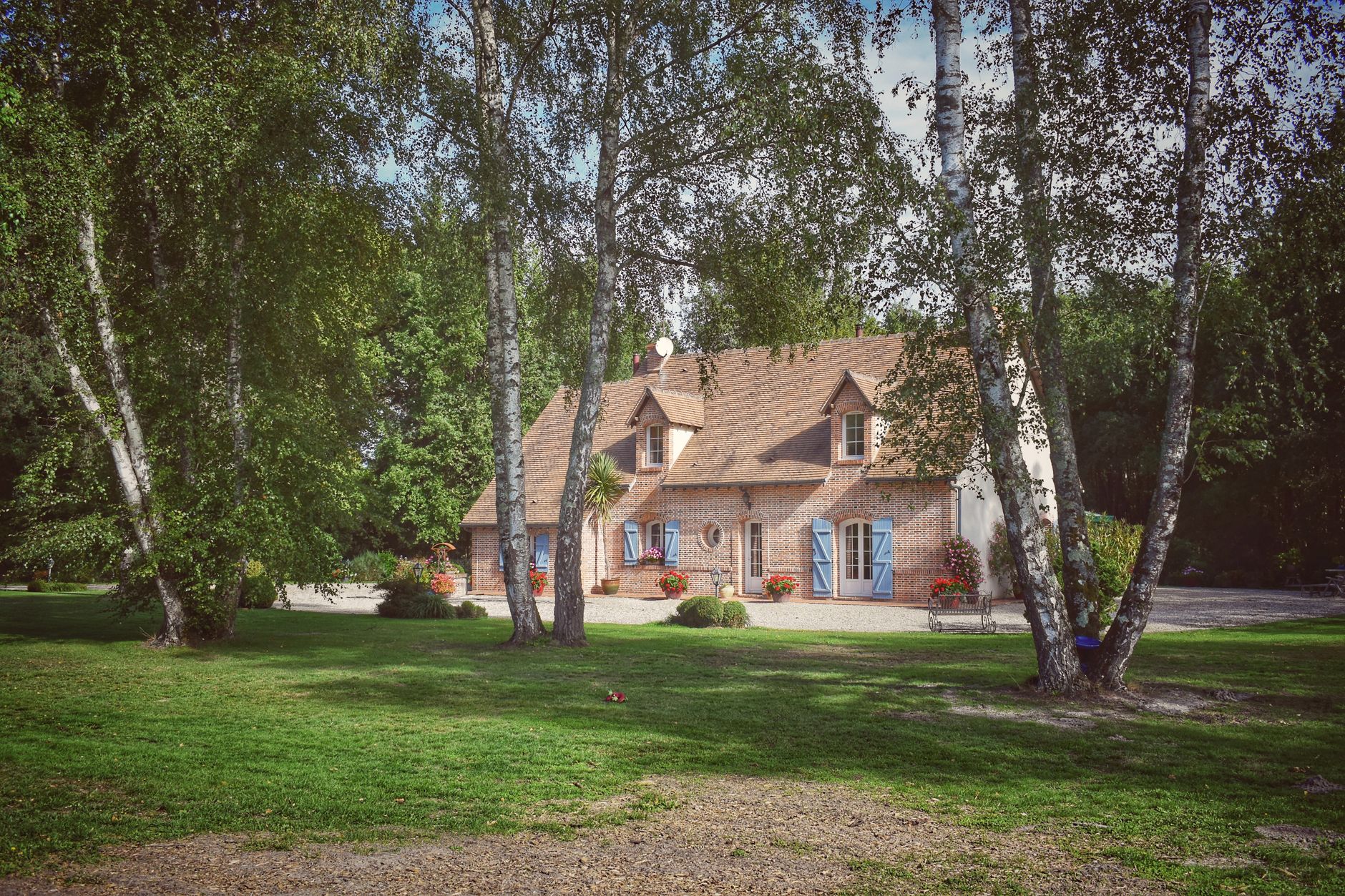 Maison de campagne en brique, volets bleus, jardin verdoyant, arbres, ciel partiellement nuageux.