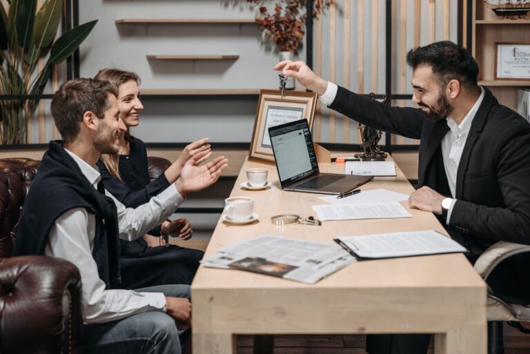 Un homme en costume tend une clé à deux personnes assises sur un canapé en cuir dans un bureau, près d'une table en bois avec ordinateur portable, tasses de café et documents.