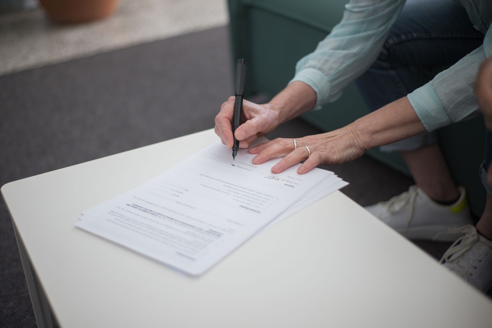 Gros plan sur des mains tenant un stylo noir au-dessus de documents papier posés sur une table blanche, avec une personne assise à côté en jean et baskets blanches.