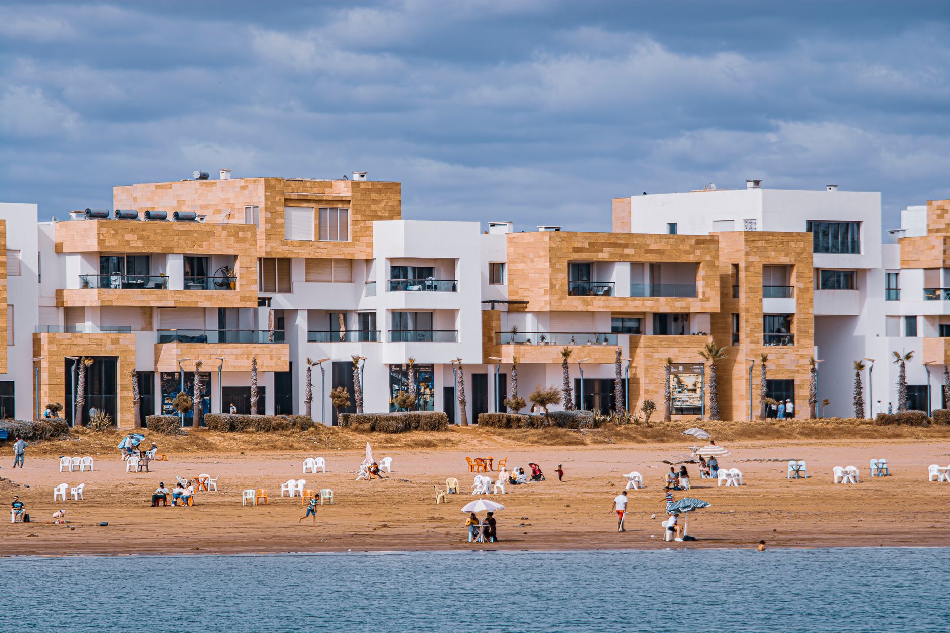 Plage avec parasols, chaises longues, bâtiments modernes en pierre claire et blancs, ciel nuageux.