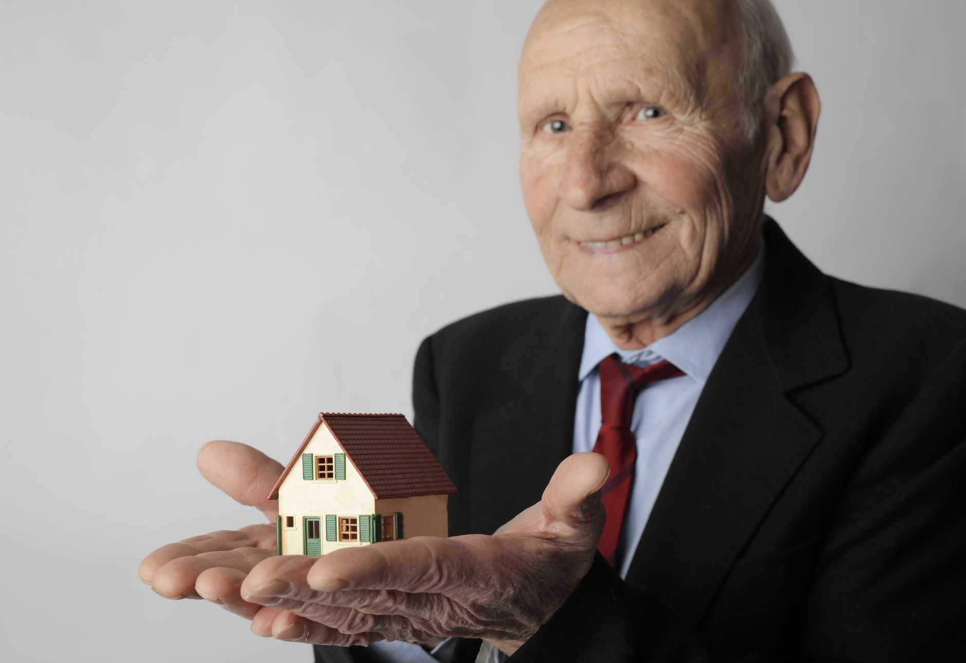 Un homme âgé en costume tient une petite maison miniature dans sa main, souriant.