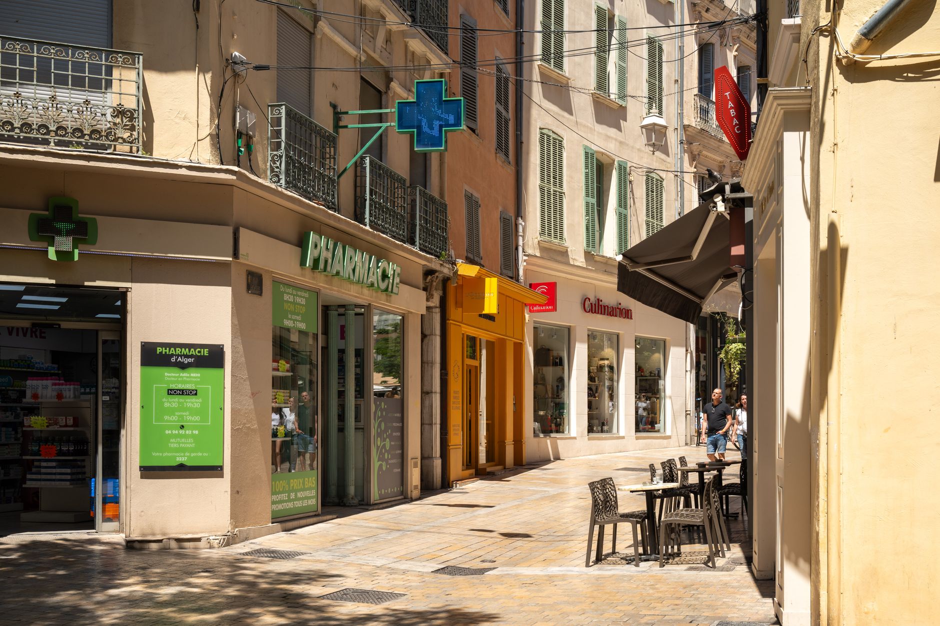 Rue piétonne animée avec pharmacie, magasin de cuisine et terrasses de café sous un soleil vif.