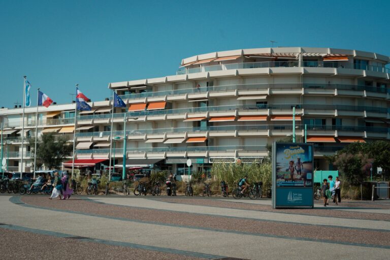 Immeuble avec balcons et stores sur promenade, vélos, personnes, drapeaux français et européens, affiche touristique.