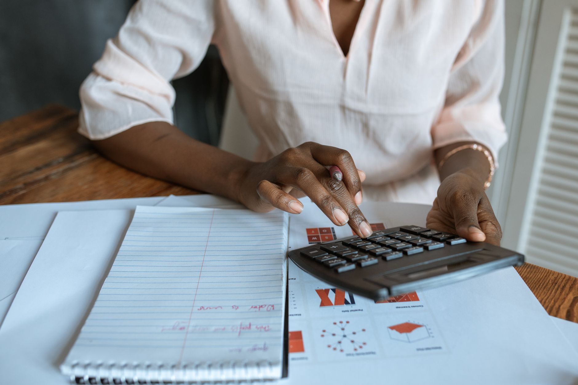Mains utilisant une calculatrice sur des documents à graphiques, à côté d’un bloc-notes à spirale, sur une table en bois.