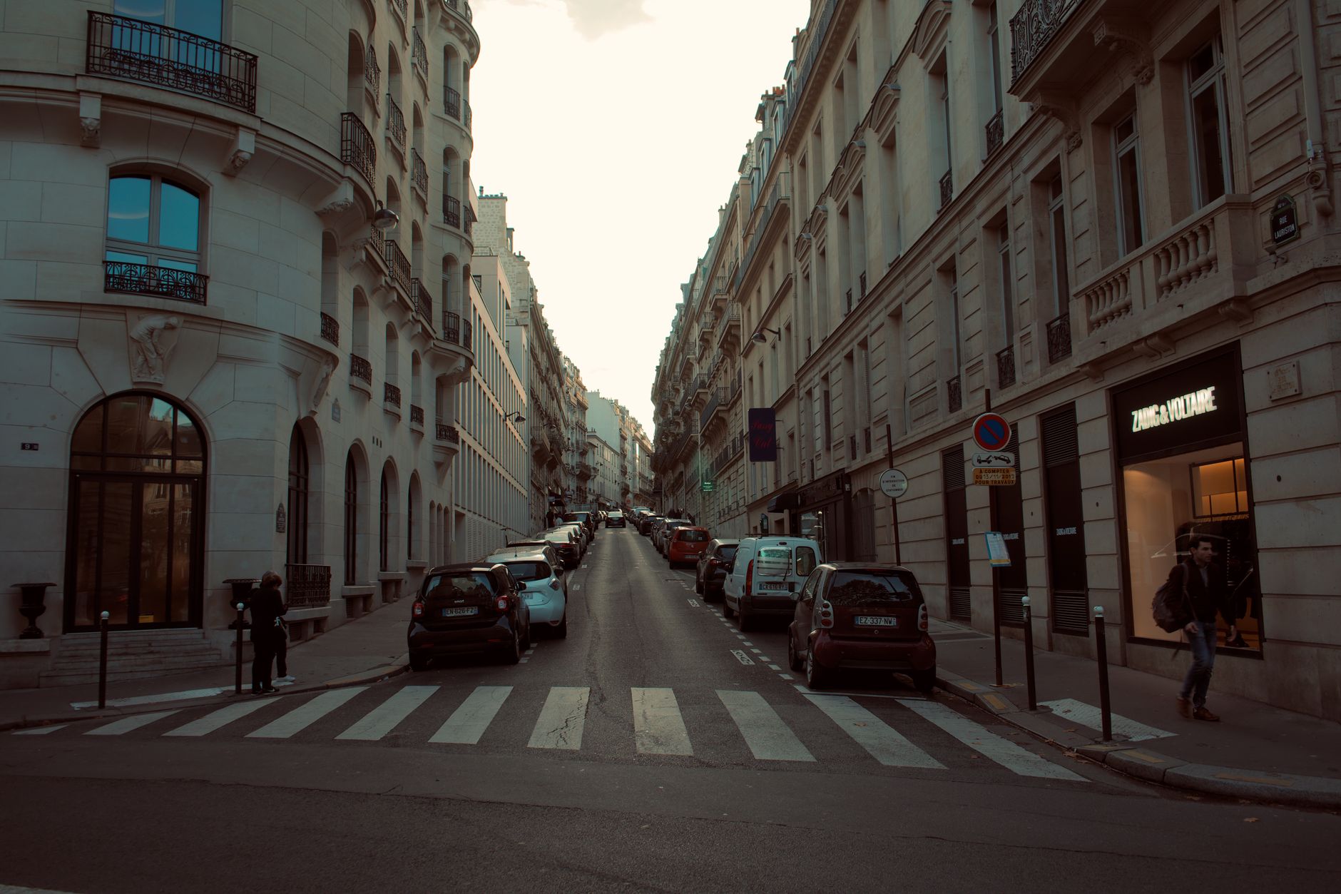 Rue bordée d’immeubles en pierre claire avec voitures garées, passage piéton et deux piétons sur les trottoirs.